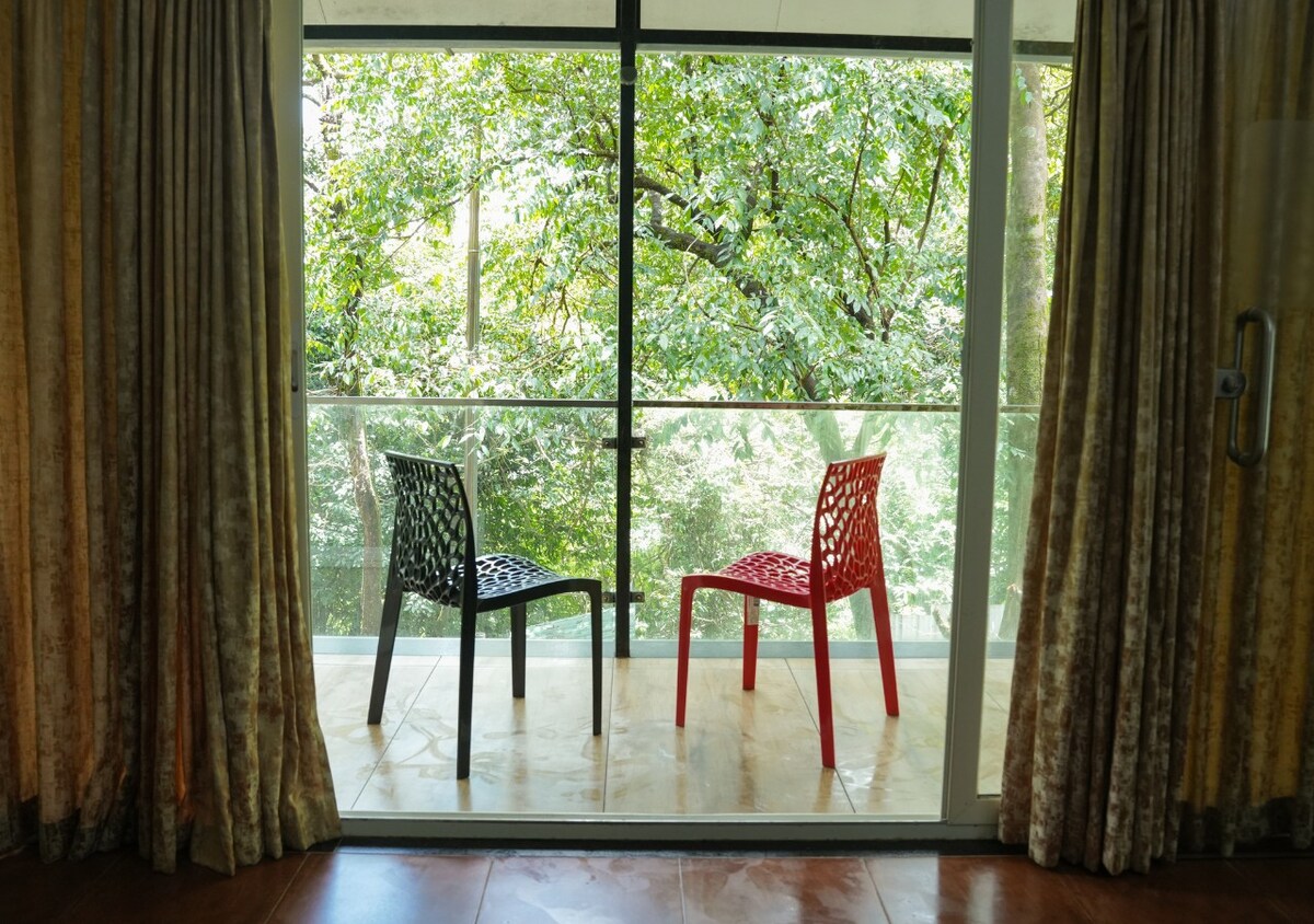 Two chairs, one black and one red, are placed on a balcony overlooking lush greenery. Glass doors provide access to the space, which is framed by soft, neutral-toned curtains, allowing natural light to brighten the area.