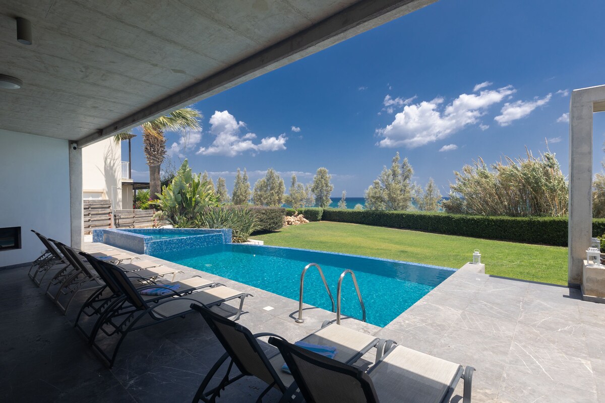 A spacious outdoor area features a private swimming pool framed by a well-maintained garden. Lounge chairs are arranged along the edge of the pool, offering a view of the sea and nearby greenery. The sky displays a blend of clouds and bright blue hues.