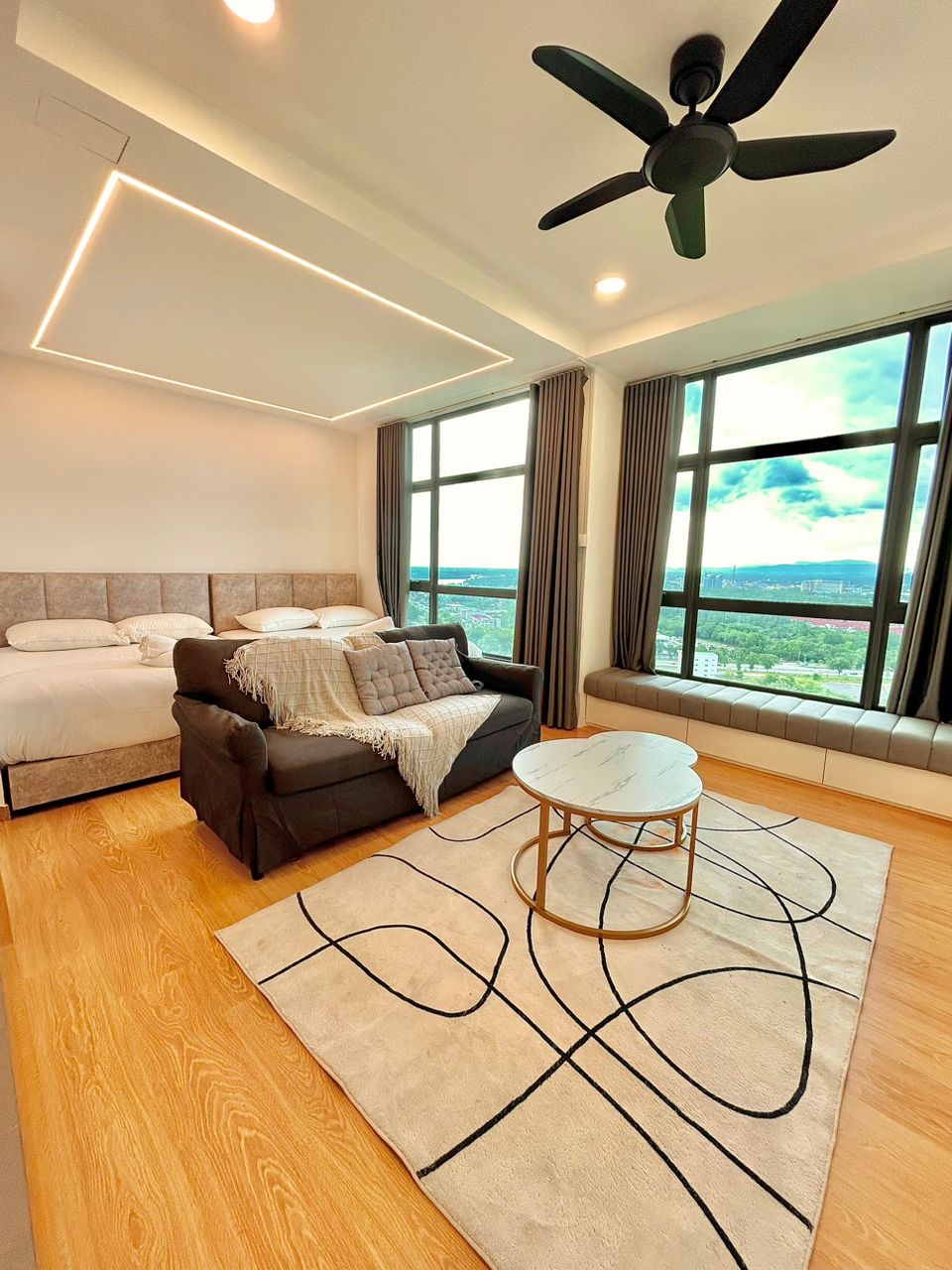 A well-lit room features large windows that offer views of the surrounding area. A spacious bed sits beside a cozy sofa, layered with a soft throw. A round table rests on a patterned rug, completing a warm and inviting atmosphere.