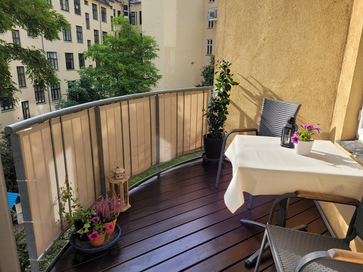 A small balcony is presented with a wooden floor. A table is set for two, accompanied by two chairs. Potted plants with flowers decorate the space, along with a lantern providing a cozy touch. A soft curtain partially shields the area from the surroundings.