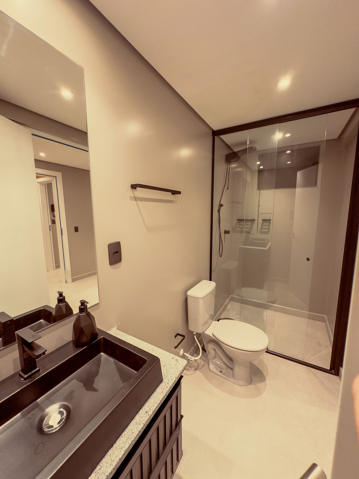 A modern bathroom is depicted featuring a sleek black sink with a granite countertop. A minimalistic shower area is enclosed in glass, and a white toilet is positioned nearby. Soft lighting illuminates the space, enhancing the neutral color palette of the walls.