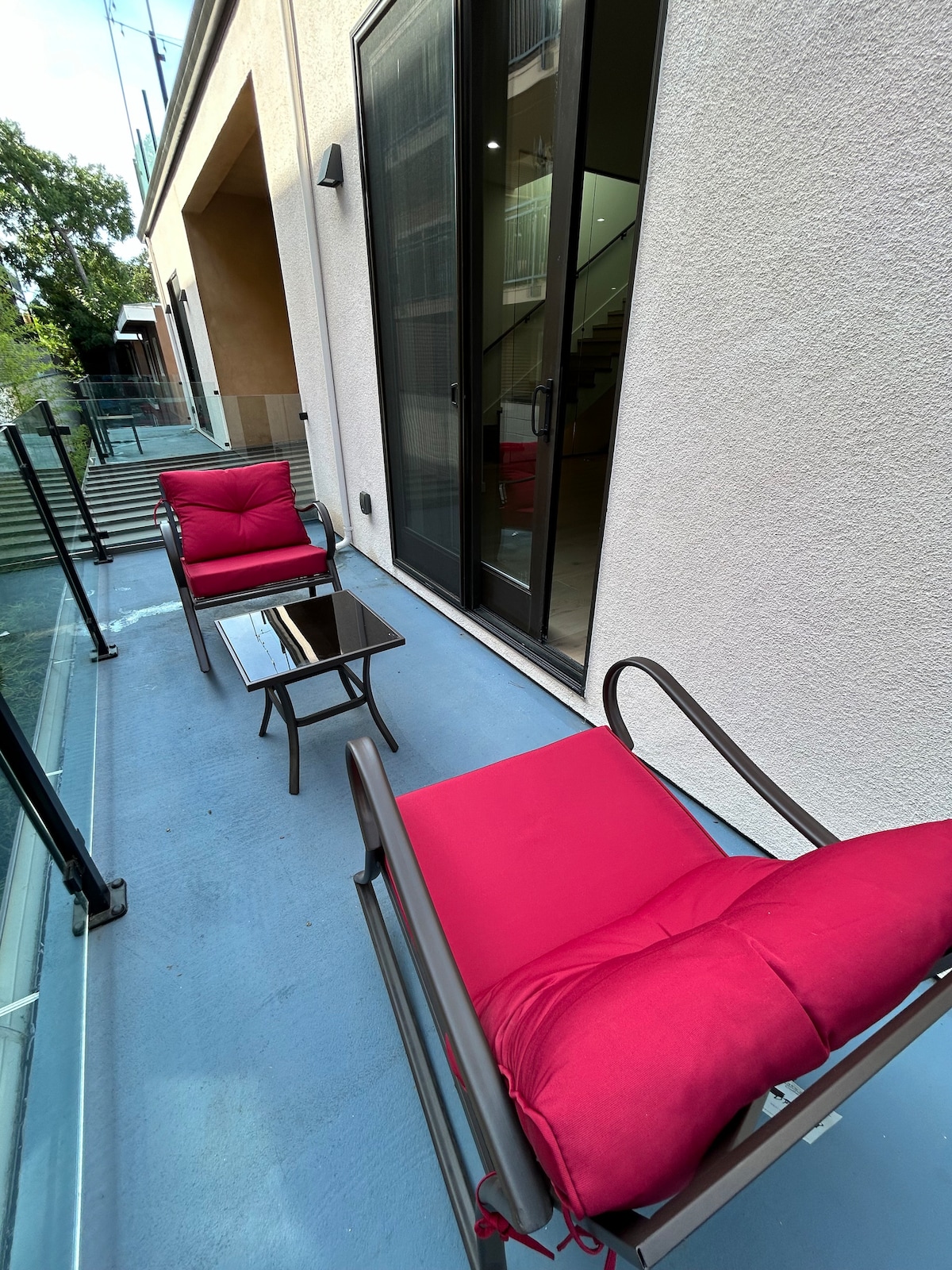A balcony area features two comfortable chairs with red cushions, positioned around a small glass-top table. The space is surrounded by sliding glass doors, providing access to the interior, and a modern staircase is visible in the background.