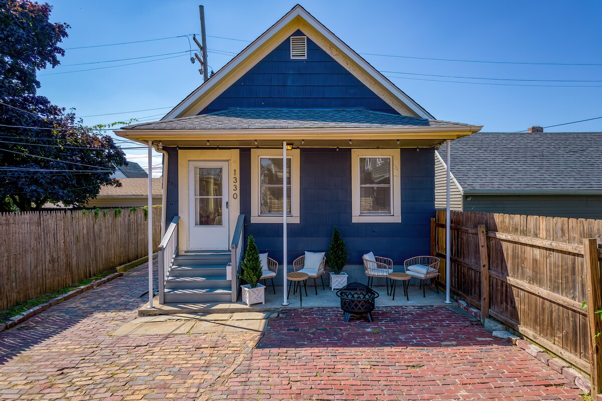 The exterior of a blue house is shown, featuring a welcoming front porch and a charming pathway of brick. Outdoor seating arrangements are visible, alongside a firepit for cozy gatherings. A fenced yard delineates the property, with clear skies overhead.