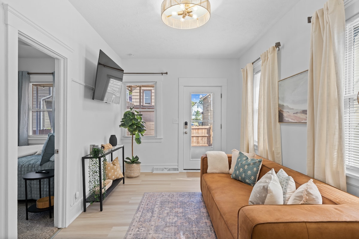 A bright living area features a warm-toned sofa adorned with decorative cushions. A modern television is mounted on the wall, while natural light fills the room through large windows with sheer curtains. A potted plant and a small shelving unit add a touch of greenery and organization.
