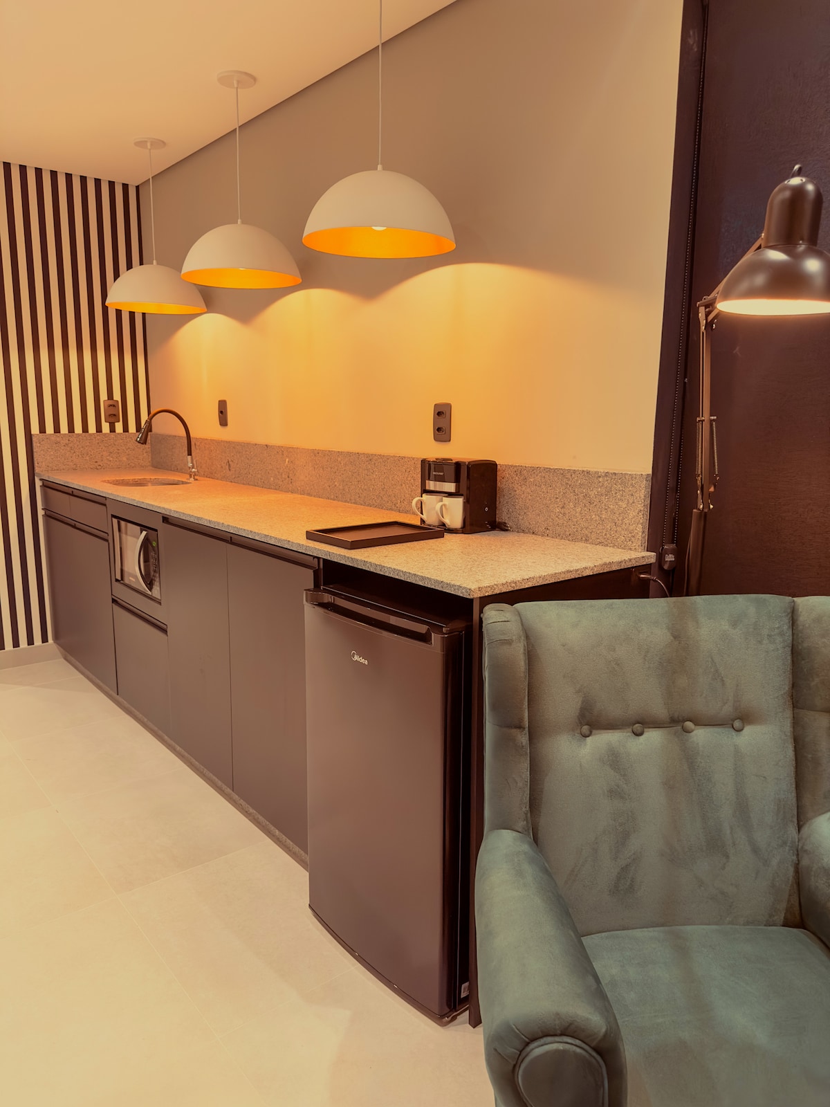 A modern kitchen area features a sleek countertop with dark cabinetry, complemented by three pendant lights casting a warm glow. Essential appliances, including a refrigerator and coffee maker, are neatly arranged, while a comfortable green armchair adds a touch of relaxation in the space.