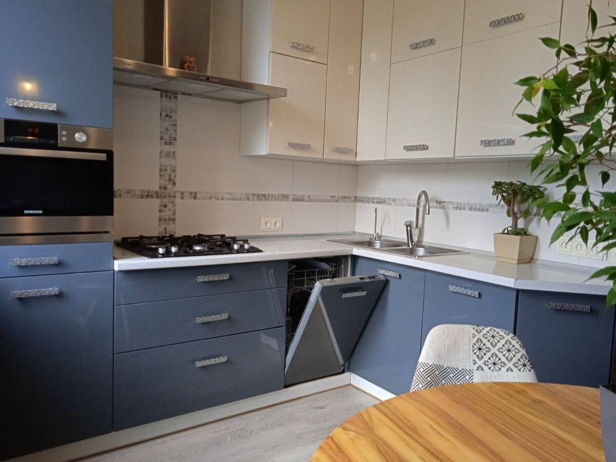 A modern kitchen is equipped with sleek gray cabinetry and stainless steel appliances. A gas stovetop is situated beside an oven, while a dishwasher is integrated within the cabinetry. A small dining table is visible, accompanied by a patterned chair and greenery in a pot.