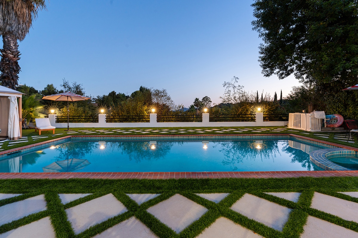 An inviting private pool is surrounded by carefully manicured grass and decorative stone pathways. Elegant lights illuminate the area, enhancing the tranquil evening ambiance. A stylish cabana and umbrella provide shaded spots for relaxation beside the pool, contributing to the outdoor escape.