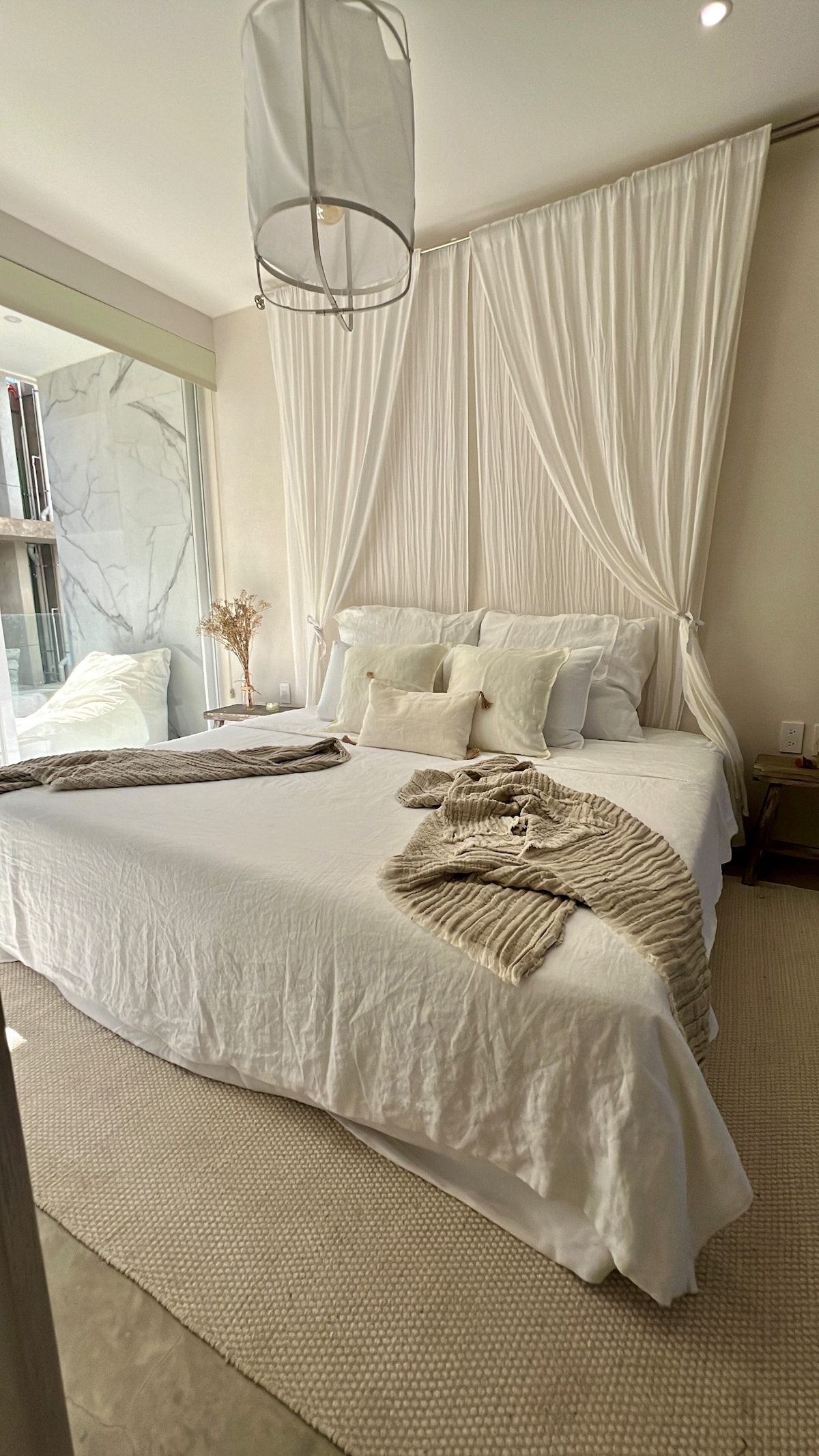 A serene bedroom is highlighted by a spacious bed dressed in crisp white linens. Soft textures are introduced through a light-colored throw and decorative pillows. Natural light floods in from a glass wall leading to a private outdoor space, creating a calming atmosphere.