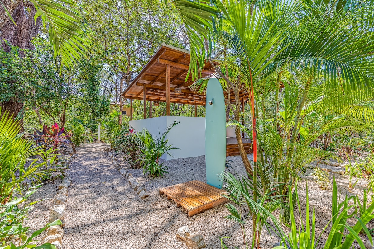 A shaded outdoor area is surrounded by lush tropical foliage and vibrant flowers. A wooden platform leads to a surfboard mounted on a white wall, creating a relaxed and inviting space perfect for enjoying nature.