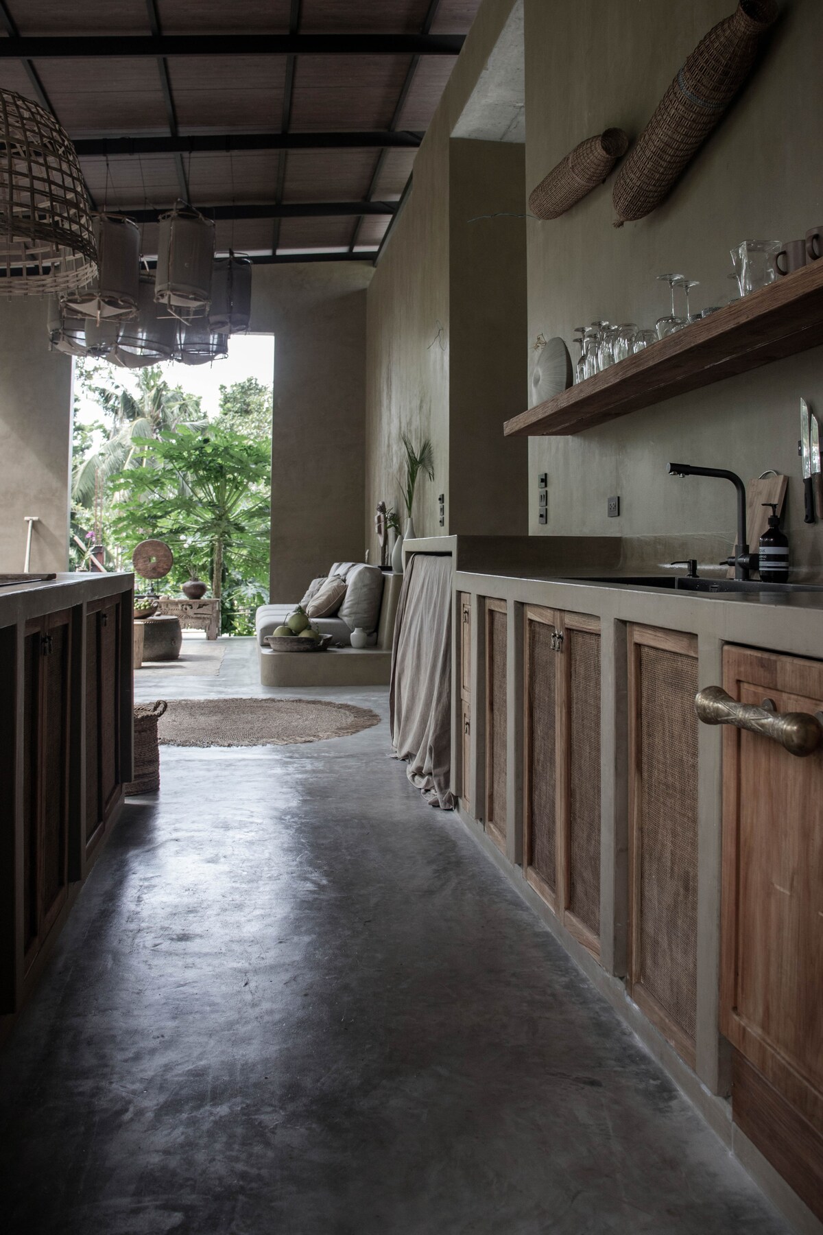 A spacious kitchen is distinguished by wooden cabinetry and a minimalist design. Natural light pours in, highlighting the earthy tones of the walls. A view of the tropical landscape is visible through open areas, while glassware rests on a shelf, enhancing the serene ambiance.