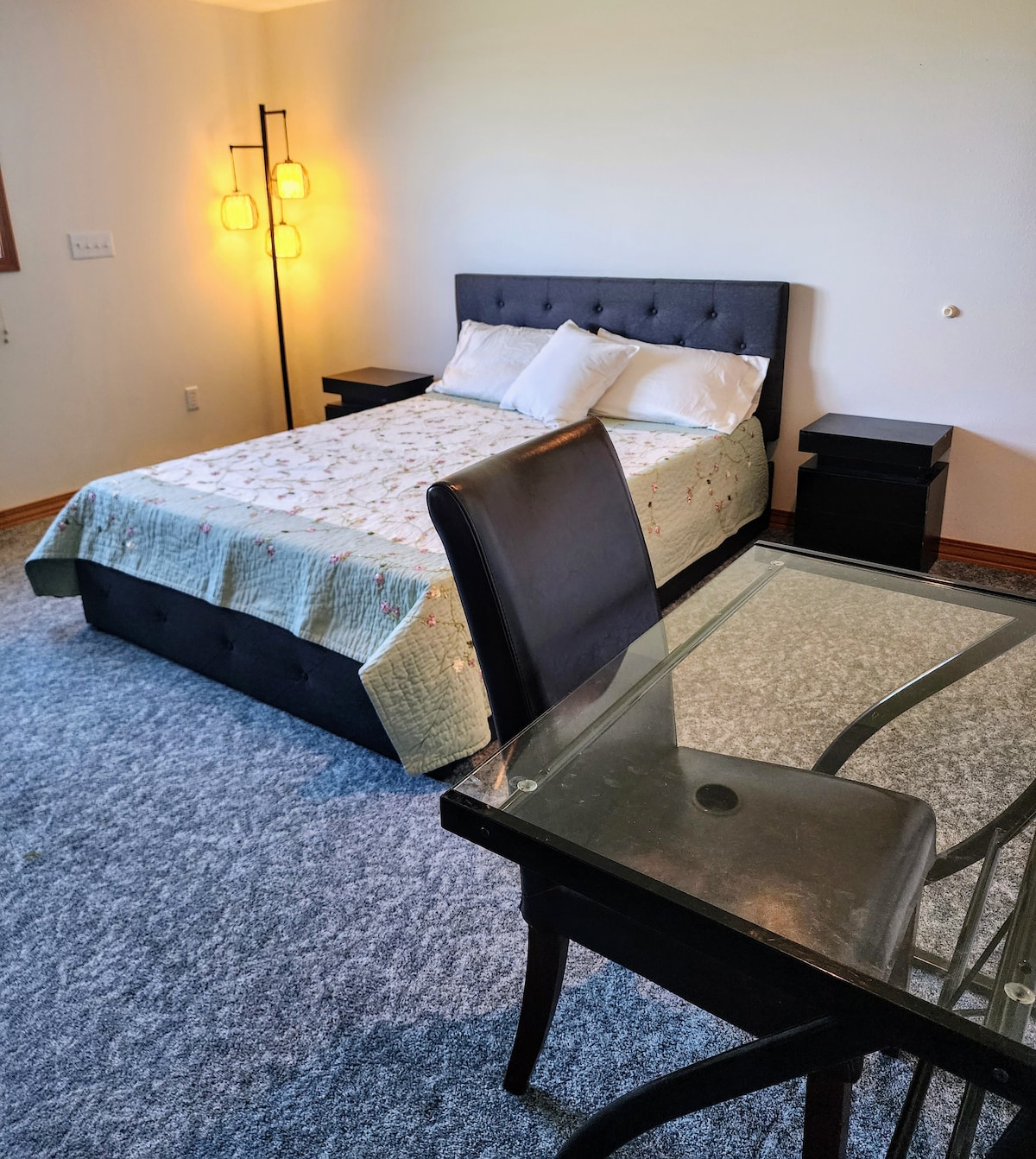 A well-lit sleeping area is furnished with a spacious bed dressed in a patterned quilt. Two nightstands frame the bed, and a small desk with a transparent top is positioned nearby. Soft carpet underfoot contributes to the room's comfort.