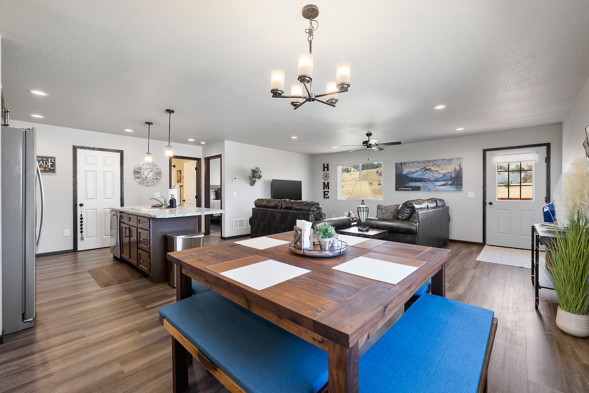 An open living space showcases a wooden dining table with blue seating, complemented by a modern kitchen featuring stainless steel appliances. A cozy lounge area is visible, furnished with dark leather sofas. Light-filled windows frame the inviting atmosphere and a mountain-themed wall is displayed in the background.