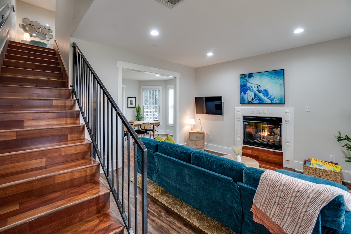 The living room features a cozy seating area with a sectional sofa in a calming blue hue. A gas fireplace is positioned beneath a wall-mounted TV, while soft lighting enhances the welcoming ambiance. A staircase with wooden steps leads to the upper level.
