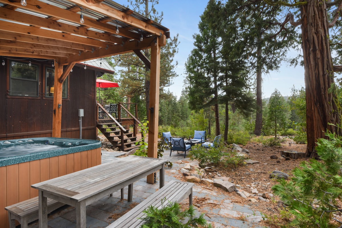 The outdoor space features a hot tub under a wooden canopy, alongside a dining table and benches. Comfortable blue chairs are positioned in the yard, surrounded by trees and natural landscaping, providing a serene ambiance for relaxation.