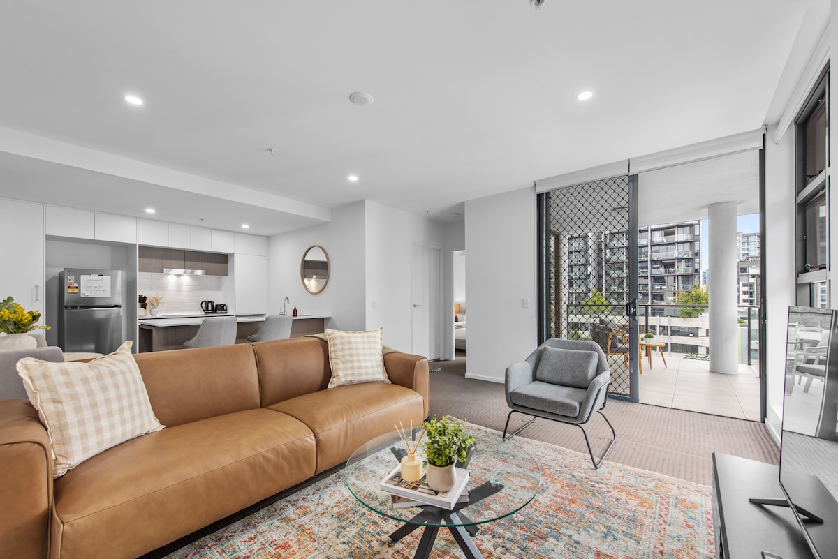 A bright living area features a brown leather sofa and a light grey armchair, providing a cozy seating arrangement. A contemporary glass coffee table sits in front, complemented by a small potted plant. Large sliding doors open to a private balcony, allowing natural light to fill the space.