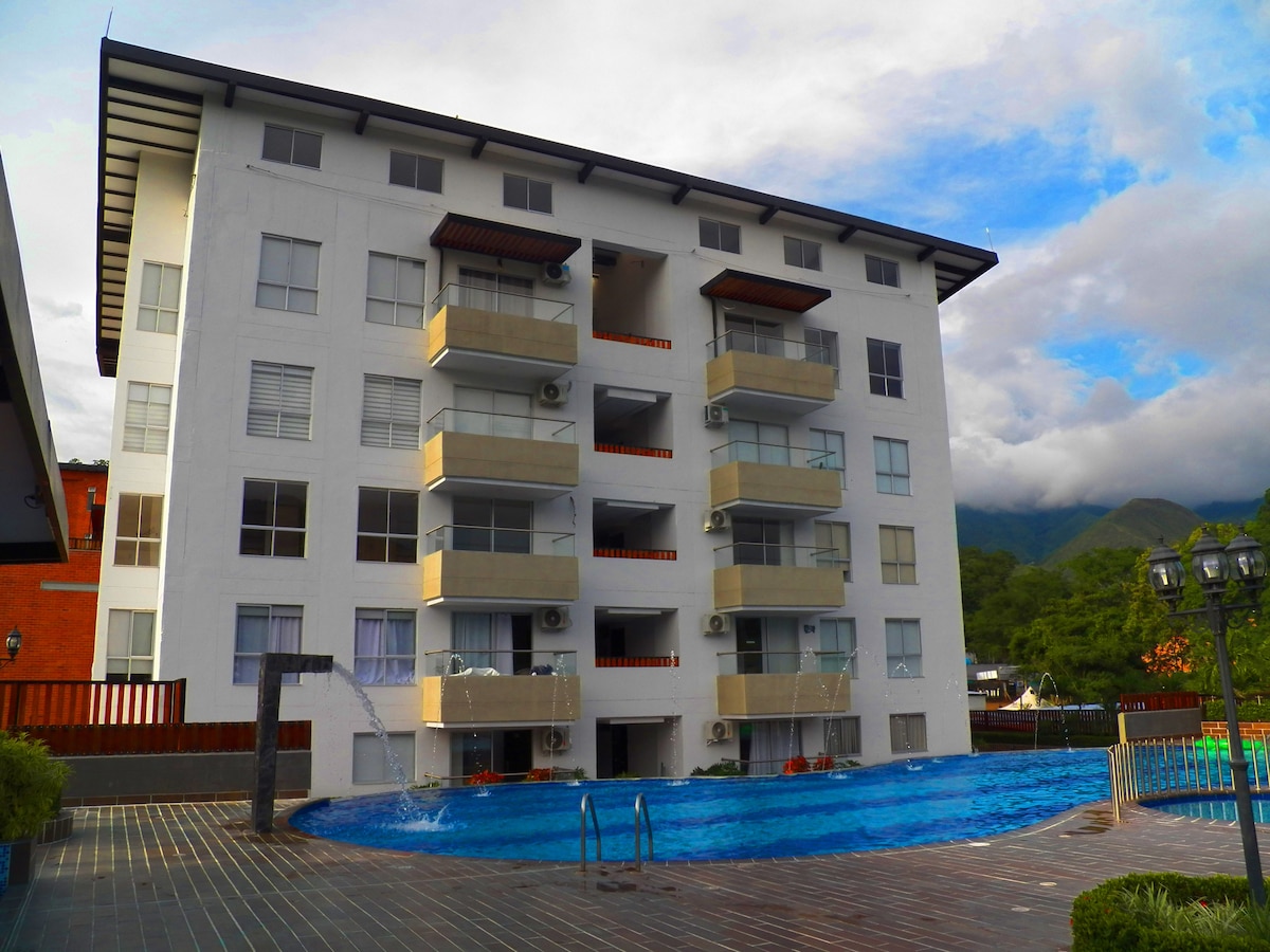 A multi-story building with a modern design features balconies on each level. The ground floor has a blue swimming pool with cascading water features. Lush greenery surrounds the property, while the backdrop highlights mountains beneath a partially cloudy sky.