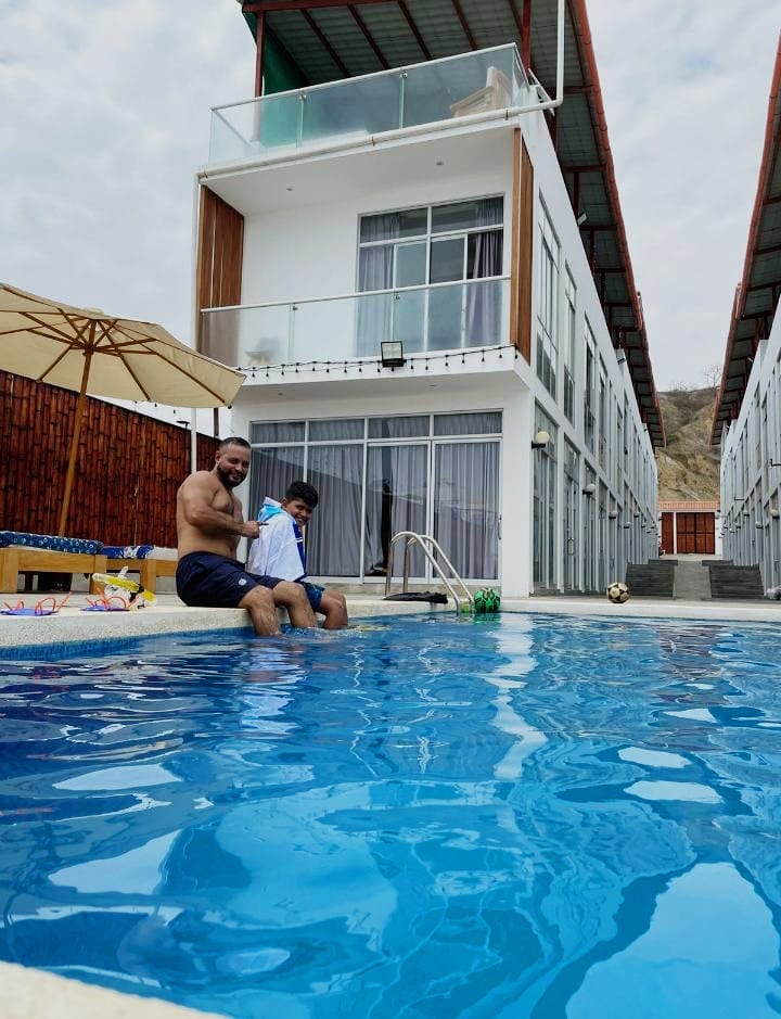 The image features a refreshing pool with clear blue water located in front of a modern multi-level building. Sun loungers and umbrellas are arranged around the pool area. Large glass doors can be seen in the background, providing ample natural light to the spacious interiors.