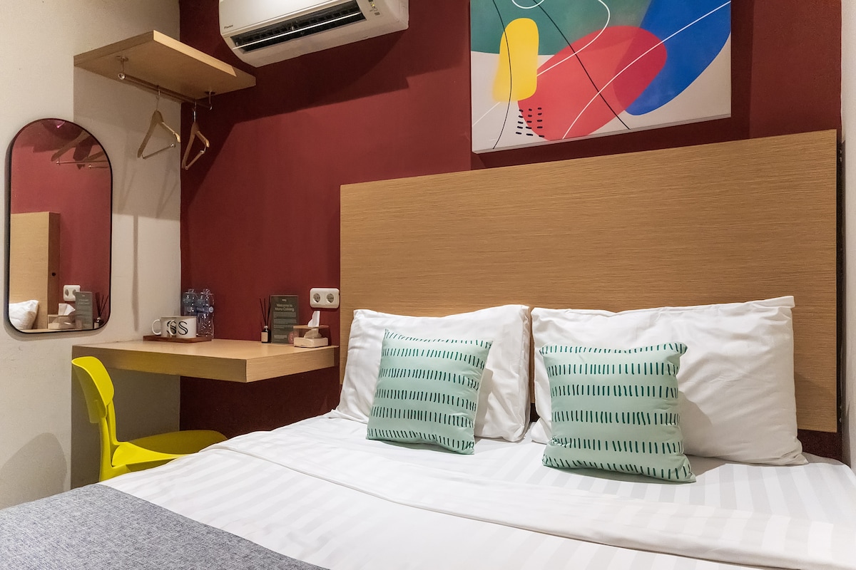 A neatly arranged sleeping area features a spacious bed with white linens and decorative cushions. A wooden headboard complements the colorful wall art above. A small desk and chair are positioned nearby, along with a mirror and coat hooks for convenience.