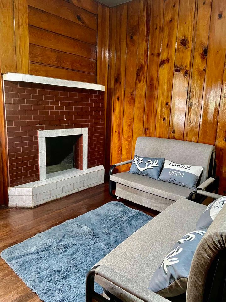 A cozy sitting area features wooden paneling and a brick fireplace. Two gray sofas with decorative pillows are positioned next to a soft area rug. Natural light enhances the warmth of the space, creating an inviting atmosphere for relaxation.