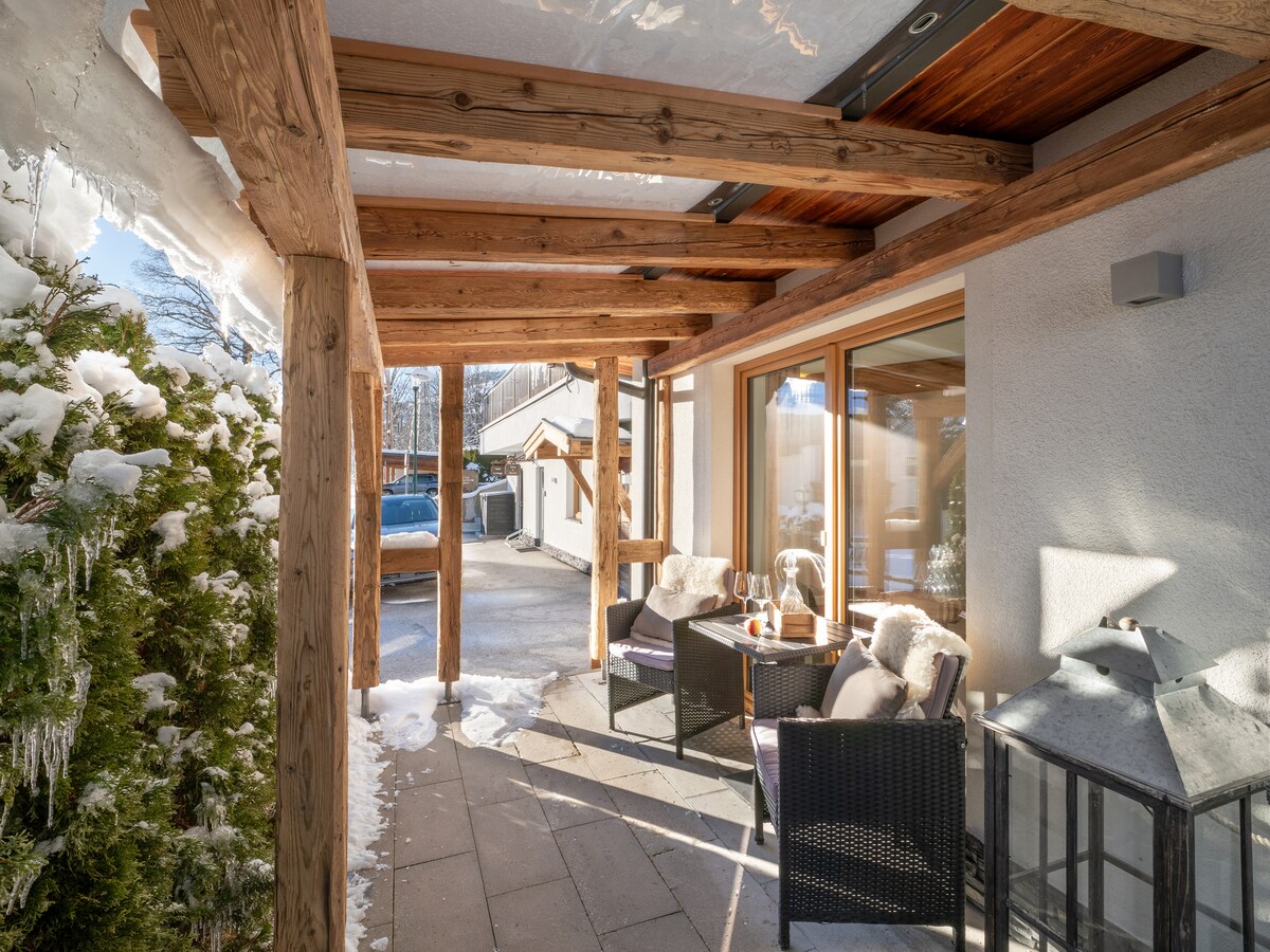 A covered outdoor terrace features a wooden framework and stone flooring, surrounded by picturesque snow-covered landscaping. Comfortable seating is arranged with a small table, inviting relaxation. Large windows allow natural light to enter the adjoining interior space.
