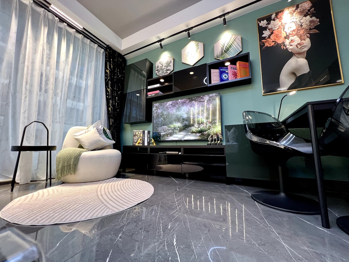 A stylish living area showcases a comfortable circular chair and a sleek black dining table with transparent chairs. A modern media console holds a large TV, framed artwork, and decorative items. Natural light filters through sheer curtains, illuminating the polished marble floor.