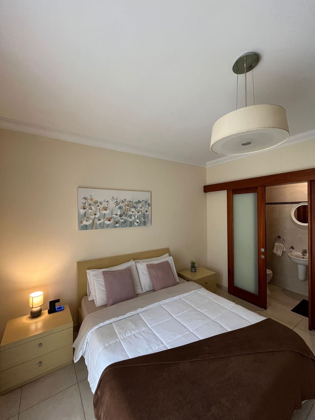 A cozy sleeping area features a spacious bed with neutral and soft-colored linens. A modern painting adorns the wall above, while bedside lamps provide gentle illumination. A sliding glass door offers access to the bathroom area, maintaining an inviting ambiance.
