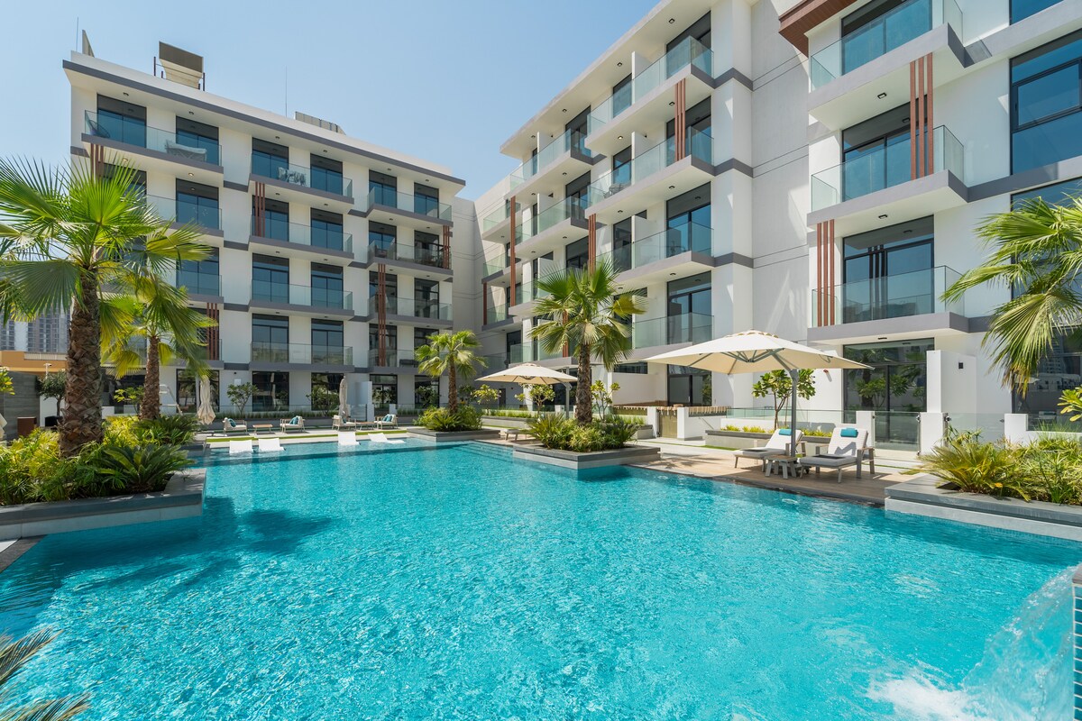 A serene pool area is surrounded by modern residential buildings. Palm trees line the edges, and sun loungers are positioned under large umbrellas near the water. Bright blue water reflects the clear sky, creating a refreshing and inviting environment.