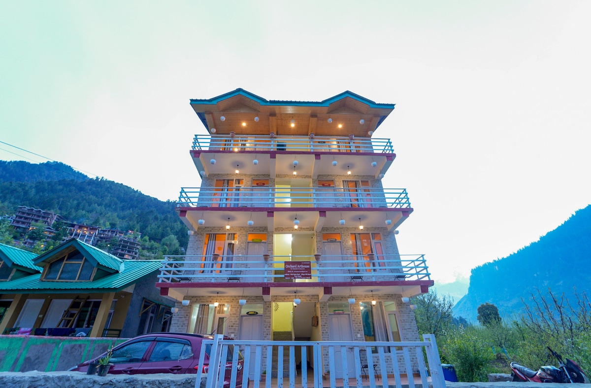 The three-story building is shown with a blend of modern architecture and natural surroundings. Multiple balconies equipped with railings enhance the exterior. Soft outdoor lighting illuminates the structure, while the backdrop features lush mountains, creating an inviting and serene setting.