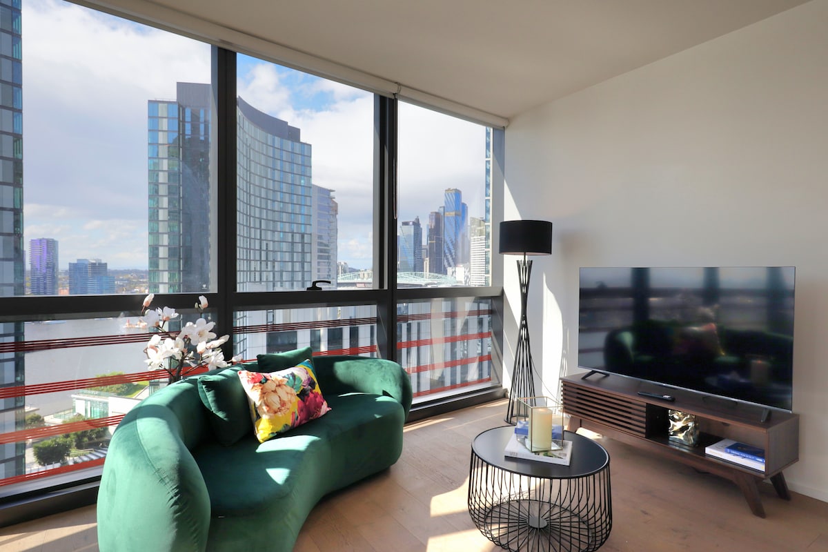 A modern living area is filled with natural light from the large panoramic windows, offering views of the city skyline. A green, curved sofa with a floral pillow is positioned near a sleek television stand, while a black lamp and a decorative table add to the contemporary design.