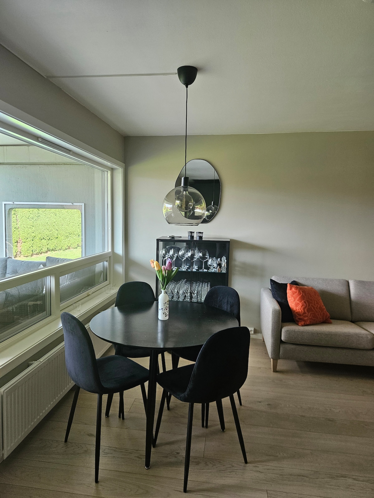 A dining area is positioned by a large window, featuring a round black table surrounded by four black chairs. A vase with fresh flowers is placed on the table. Behind the table, a gray sofa is located, complemented by a decorative orange cushion.