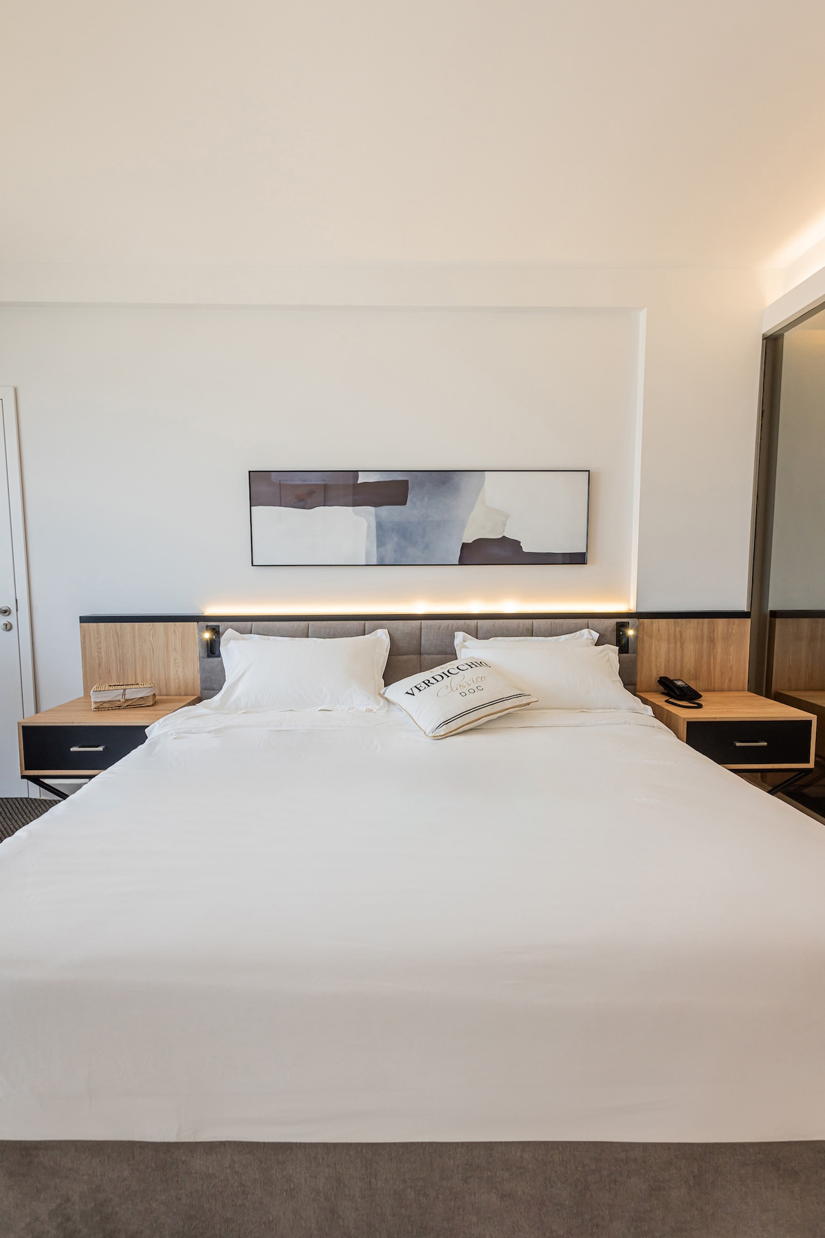 A spacious bedroom features a large bed with a crisp white duvet and decorative pillows. Soft lighting is integrated into the headboard, complementing the simple artwork above. A mirrored closet reflects the room's elegance, enhancing the overall contemporary design.