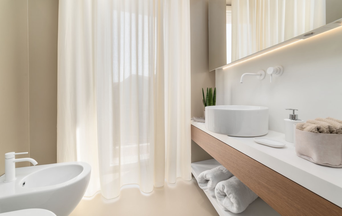 A modern bathroom is equipped with a round sink atop a light wood vanity. A bidet is positioned nearby, while soft towels are neatly arranged on a lower shelf. Natural light filters through sheer curtains, creating a serene and bright ambiance.