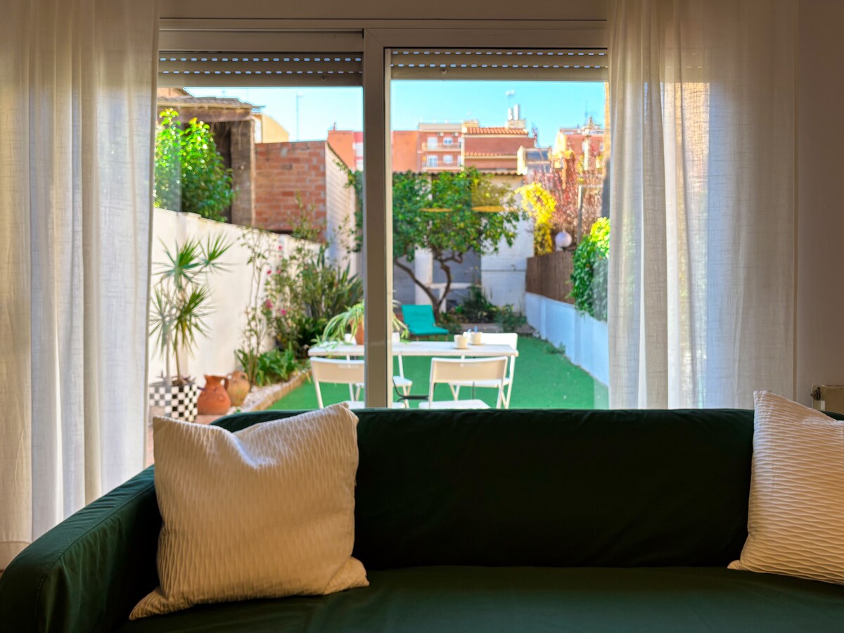 A view through a large window reveals a patio featuring green grass, seating areas, and potted plants. Sheer curtains provide a soft frame around the outdoor scene, with bright sunlight illuminating the space.