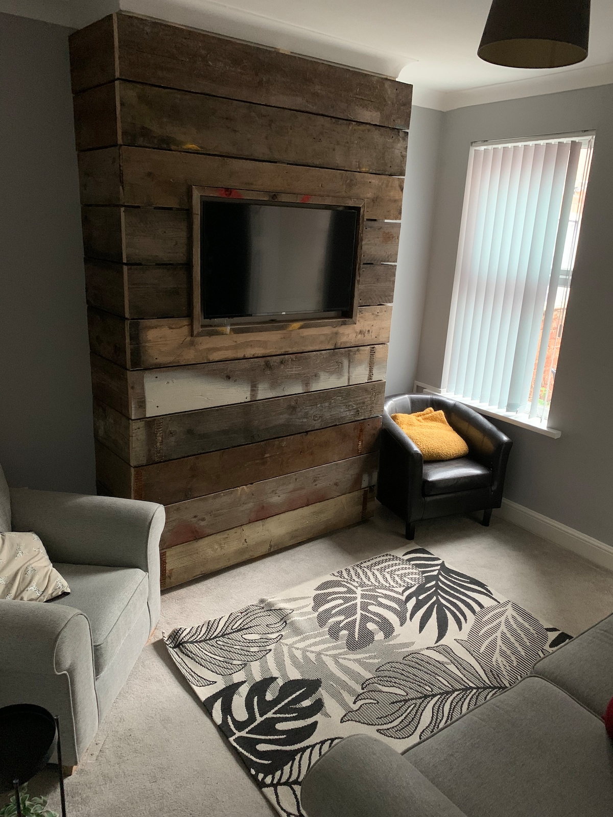 A cozy living area features a rustic wooden accent wall with a mounted flat-screen television. A comfortable armchair is positioned nearby, accompanied by a yellow cushion. Natural light enters through vertical blinds, illuminating a patterned rug that adds warmth to the space.