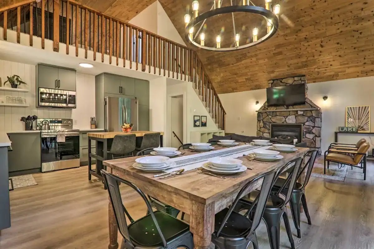 An inviting dining area features a rustic wooden table set with place settings. Above, a large circular chandelier hangs from the vaulted ceiling. The kitchen area is visible in the background, showcasing modern appliances and wooden accents, while a cozy seating area is adjacent.