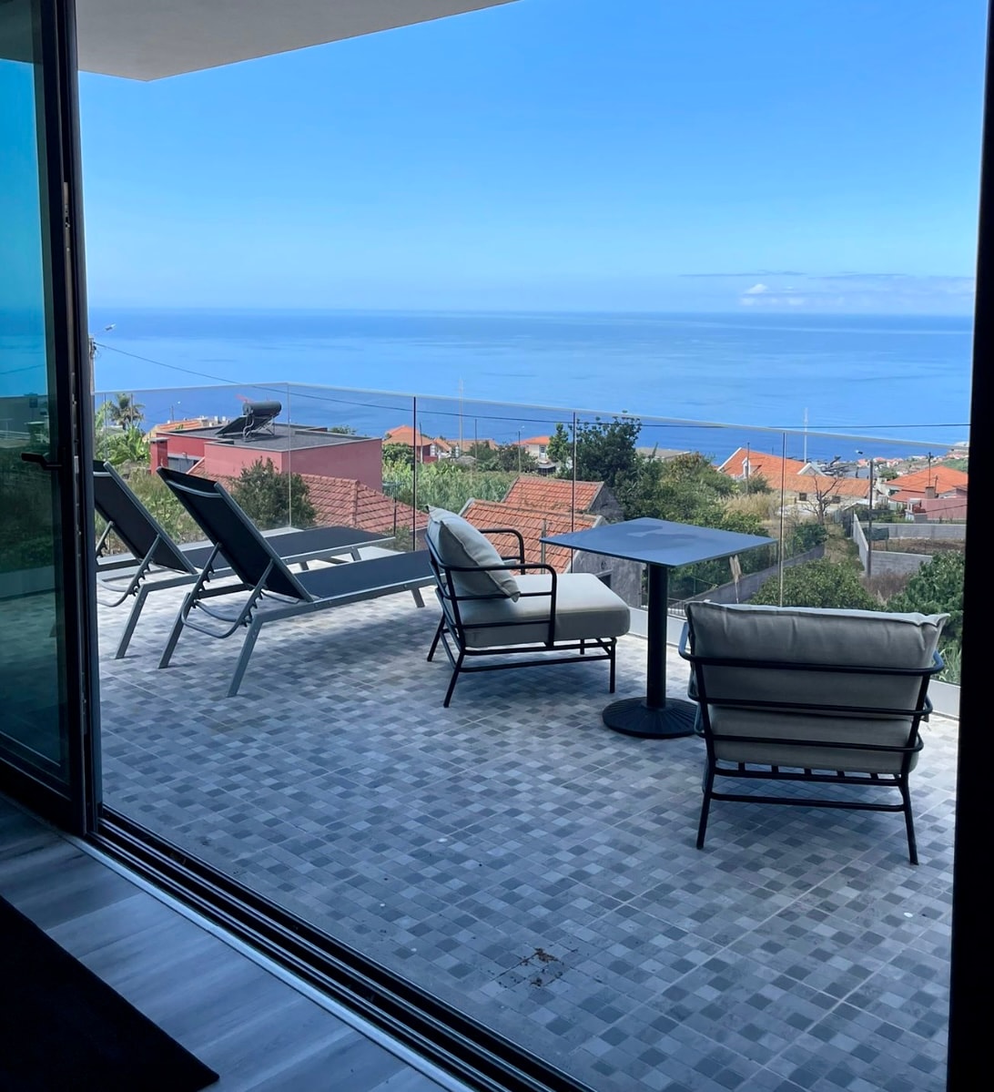 A scenic outdoor terrace is presented, featuring sleek lounge chairs and a small table. The expansive ocean view stretches beyond, complemented by distant coastal scenery. The terrace's tiled flooring enhances the modern aesthetic of the space.