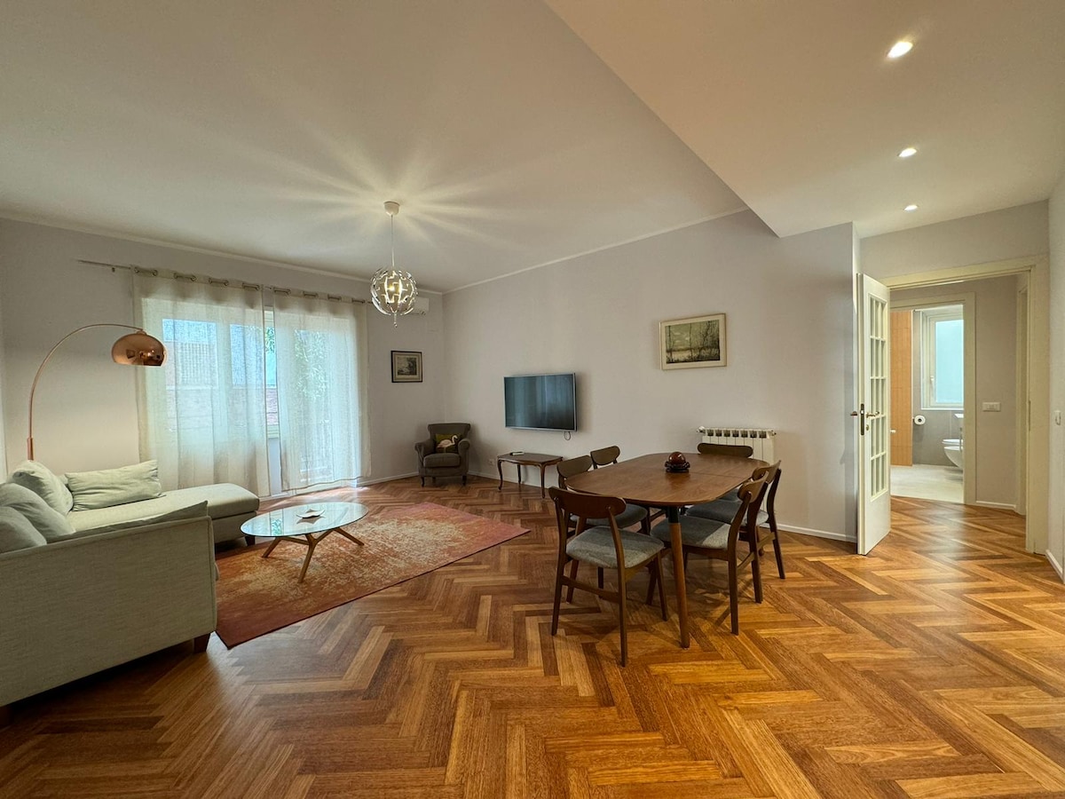 A spacious living room features a light-colored sofa and a glass coffee table, set on a wooden floor with a rug underneath. A dining table with wooden chairs is positioned nearby. Large windows allow natural light to fill the room, while a modern light fixture hangs above.