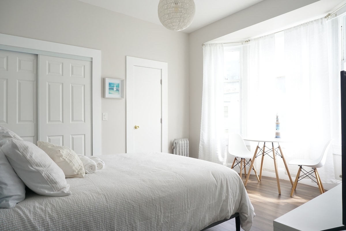 A spacious bedroom features a soft-colored quilted bed with plush pillows. Natural light floods in through large windows dressed in sheer curtains. A small table with white chairs is positioned near the window, enhancing the room's airy feel.