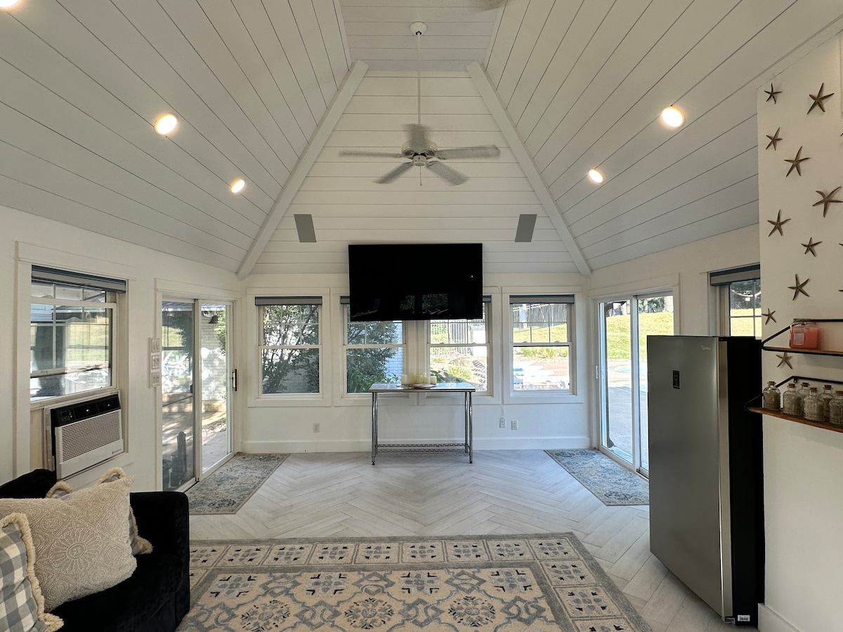 The sunroom features a high, sloped ceiling with white paneling and gentle lighting. Large windows surround the space, offering views of the outdoors. A contemporary sofa complements the room, and a television is mounted on the wall. A compact refrigerator is positioned nearby.