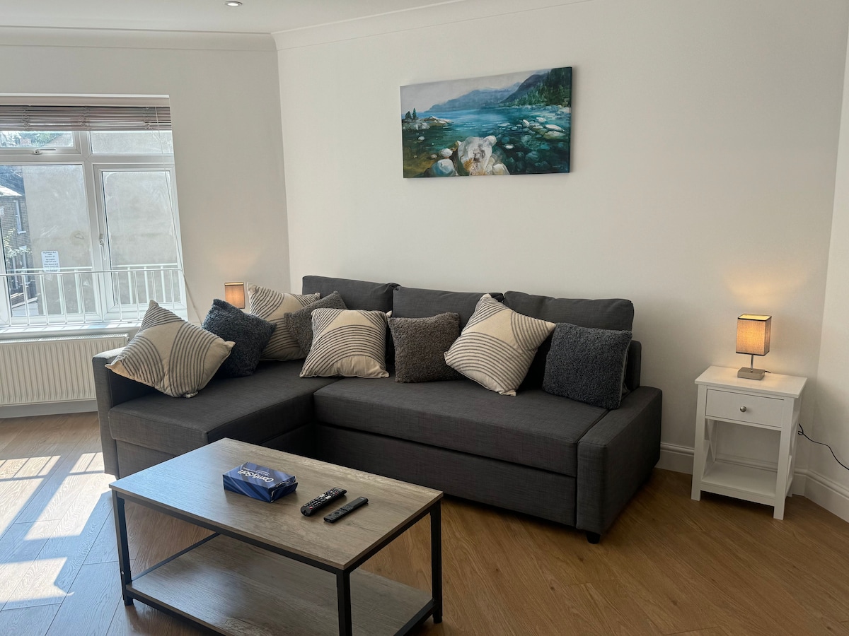 A living area showcases a dark gray sectional sofa adorned with striped and solid pillows. A small wooden coffee table is positioned in front, accompanied by a remote control. A painting of a serene landscape hangs above the sofa, with natural light streaming through the window.