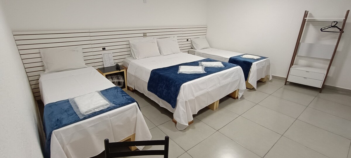 The room features two single beds and one spacious double bed, all dressed in white linens with blue accents. A simple wooden chair and a minimalist shelving unit are included. Natural light brightens the neutral-colored walls and floor tiles.