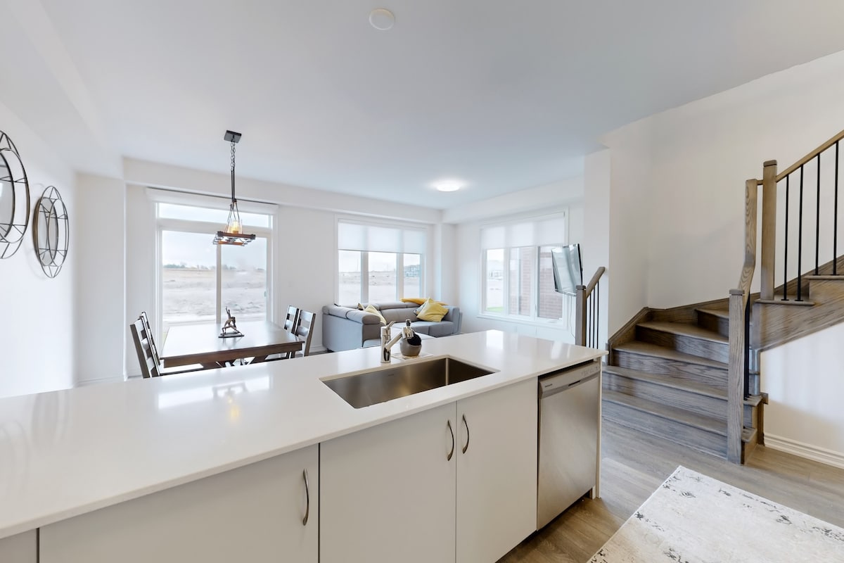 A well-equipped kitchen features modern cabinetry and a sleek countertop. Visible is a dining area with a wooden table and four chairs, and a cozy living space with a sectional sofa adorned with cushions. Natural light flows in through large windows, enhancing the bright ambiance.
