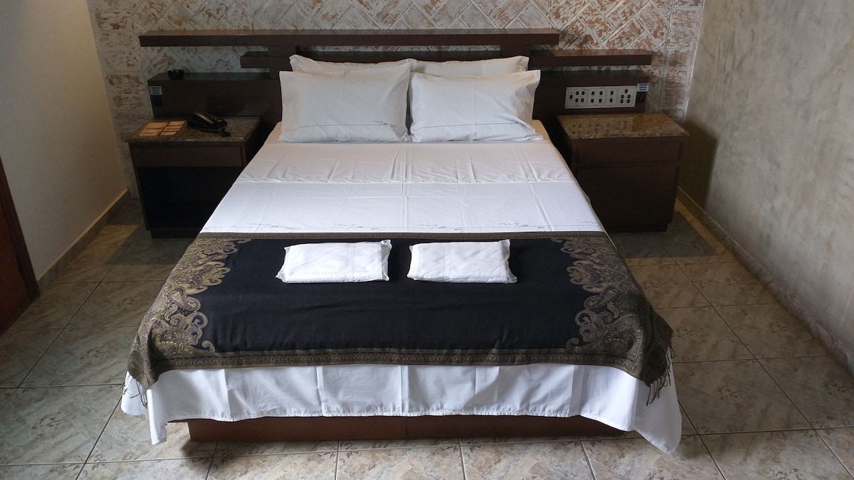 A neatly arranged sleeping area features a spacious bed with white linens and a decorative black runner. Two folded towels are placed at the foot of the bed. The headboard is minimalistic, complemented by bedside tables with lamps, on a tiled floor.