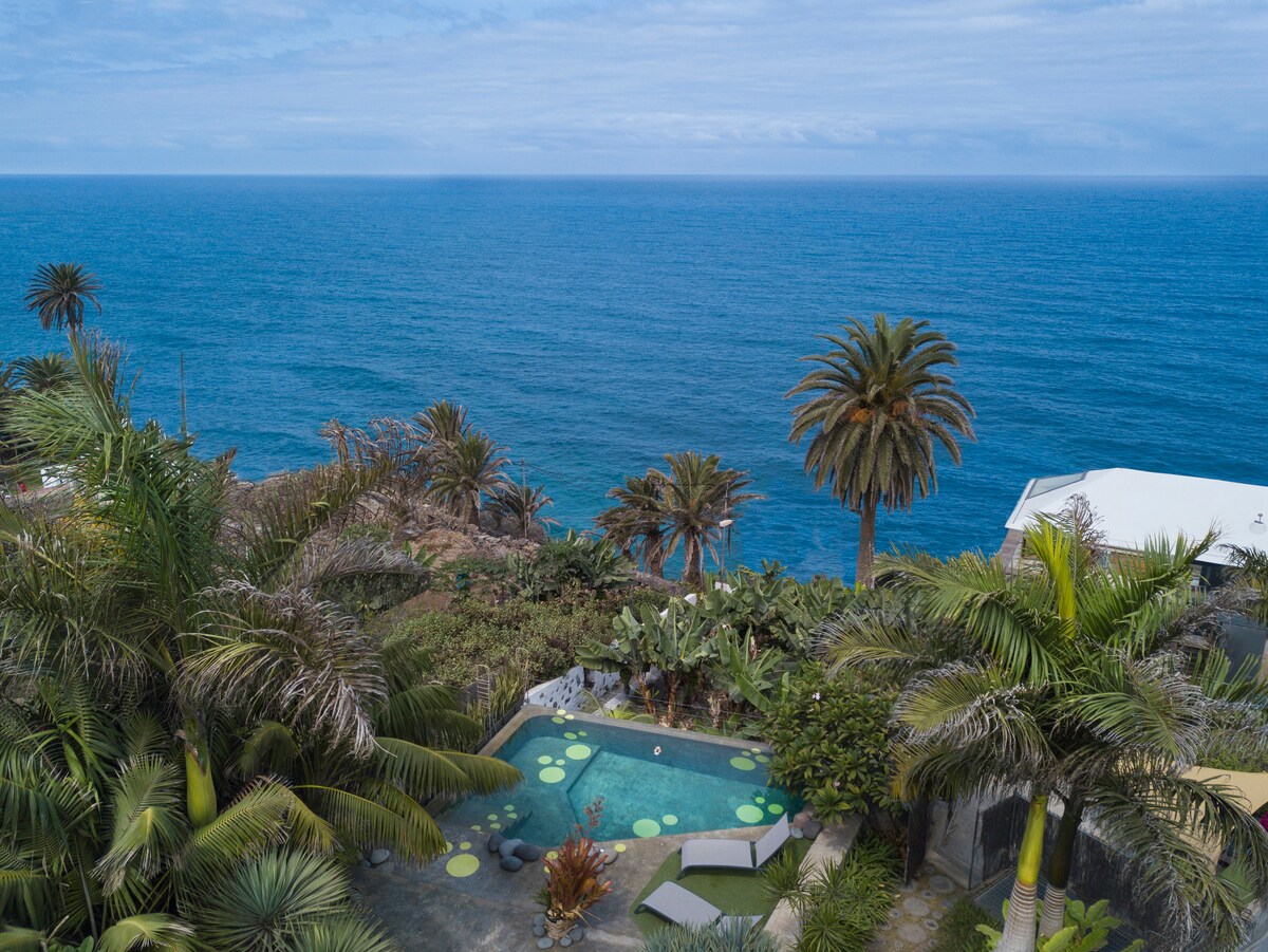 An expansive view of the ocean is framed by lush palm trees and tropical vegetation. A serene swimming pool, surrounded by green accents and lounge chairs, provides a tranquil space. The soft blue of the sea blends seamlessly with the sky on the horizon.