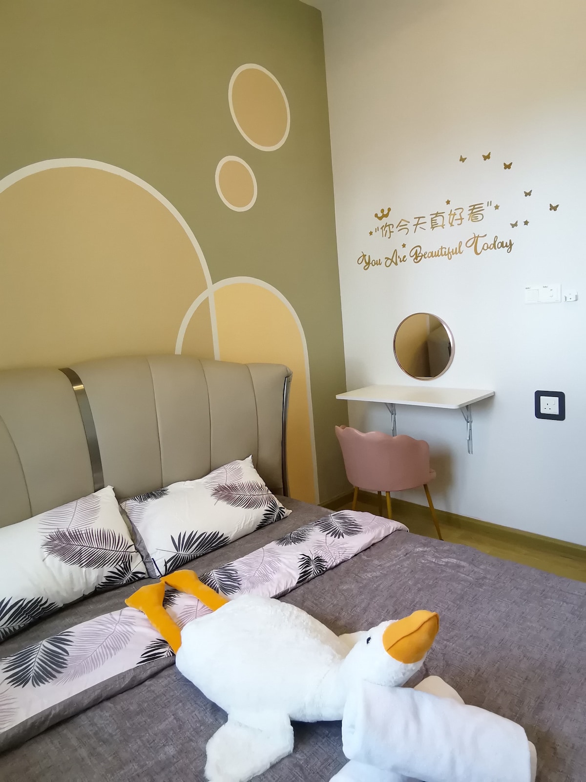 A cozy sleeping area features a spacious bed with decorative pillows, alongside a wall adorned with soft green hues and circular designs. A compact vanity with a round mirror and a pink chair complements the space, while a plush duck toy adds a playful touch.