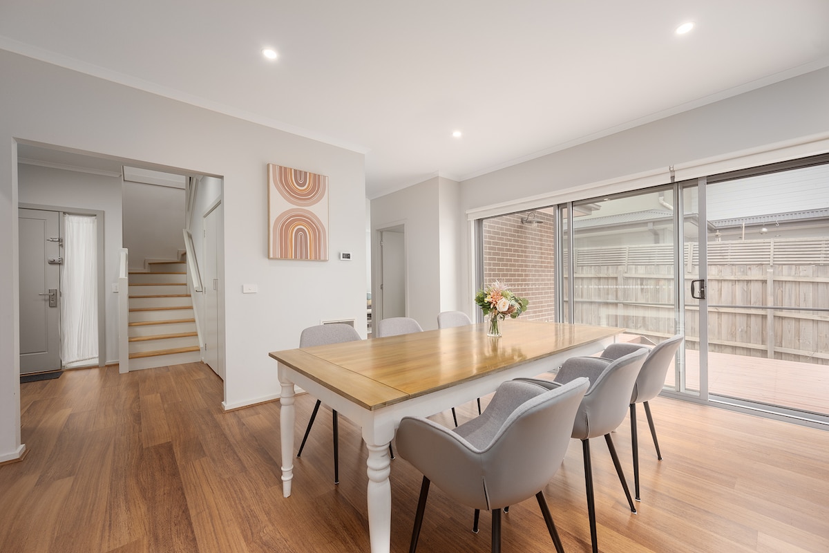 The dining area features a large wooden table surrounded by six upholstered chairs, offering ample seating for meals. Large glass sliding doors provide natural light and an unobstructed view of the private backyard, while warm wood flooring enhances the inviting ambiance.