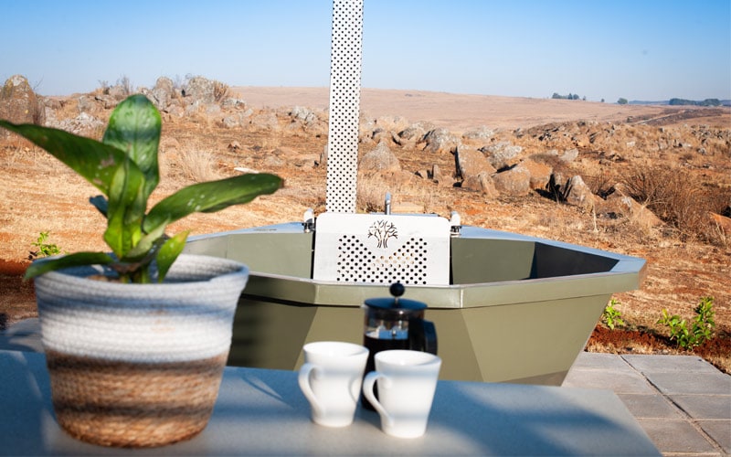 An outdoor wood-fired hot tub provides a serene retreat, set against a vast, open landscape of rolling hills. A potted plant and two white cups are positioned on a table, adding a touch of comfort to the setting.