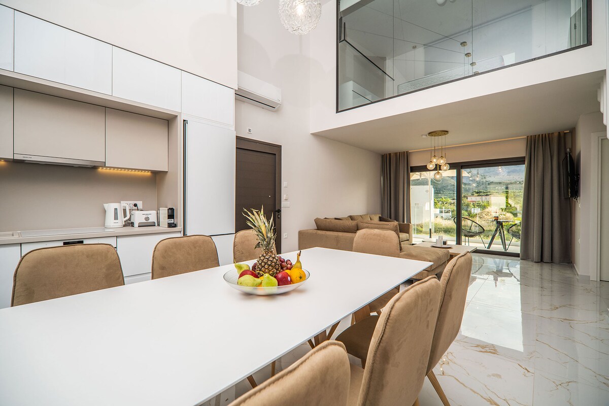 A modern dining area features a spacious white table surrounded by comfortable beige chairs. A bowl of fresh fruit is centered on the table, while large windows allow natural light to illuminate the room. Views of the outside landscape can be seen through the glass doors.