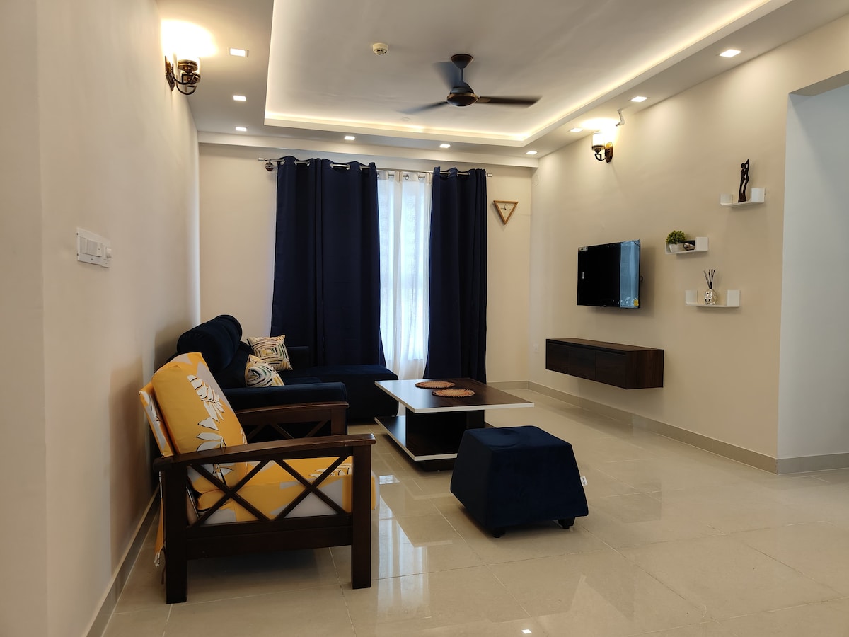 The living room features a comfortable seating arrangement, including a dark blue sofa and an armchair with a bright yellow cushion. A coffee table sits at the center, complemented by a television mounted on the wall. Soft lighting enhances the airy space.