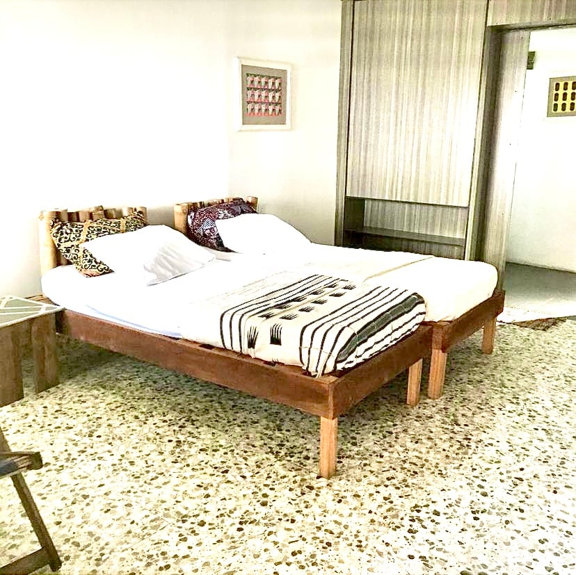 A spacious room features two beds with neutral linens and decorative throw pillows. A textured wall adds visual interest. The floor showcases a smooth, speckled finish, complementing the overall room design. Soft light enters through an adjacent door, enhancing the inviting ambiance.